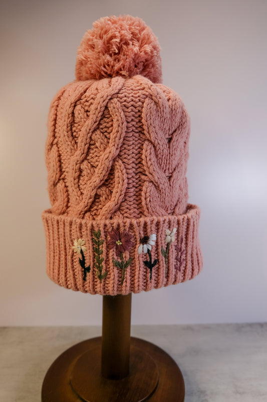Knitted pink beanie with embroidered flowers on a wooden stand against a plain background