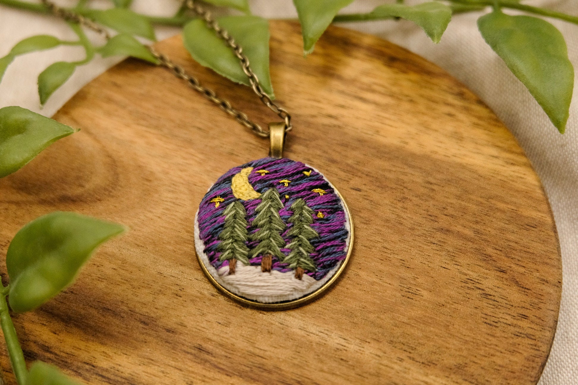 Embroidered bronze pendant featuring evergreen trees, a night sky with stars, moon and snow