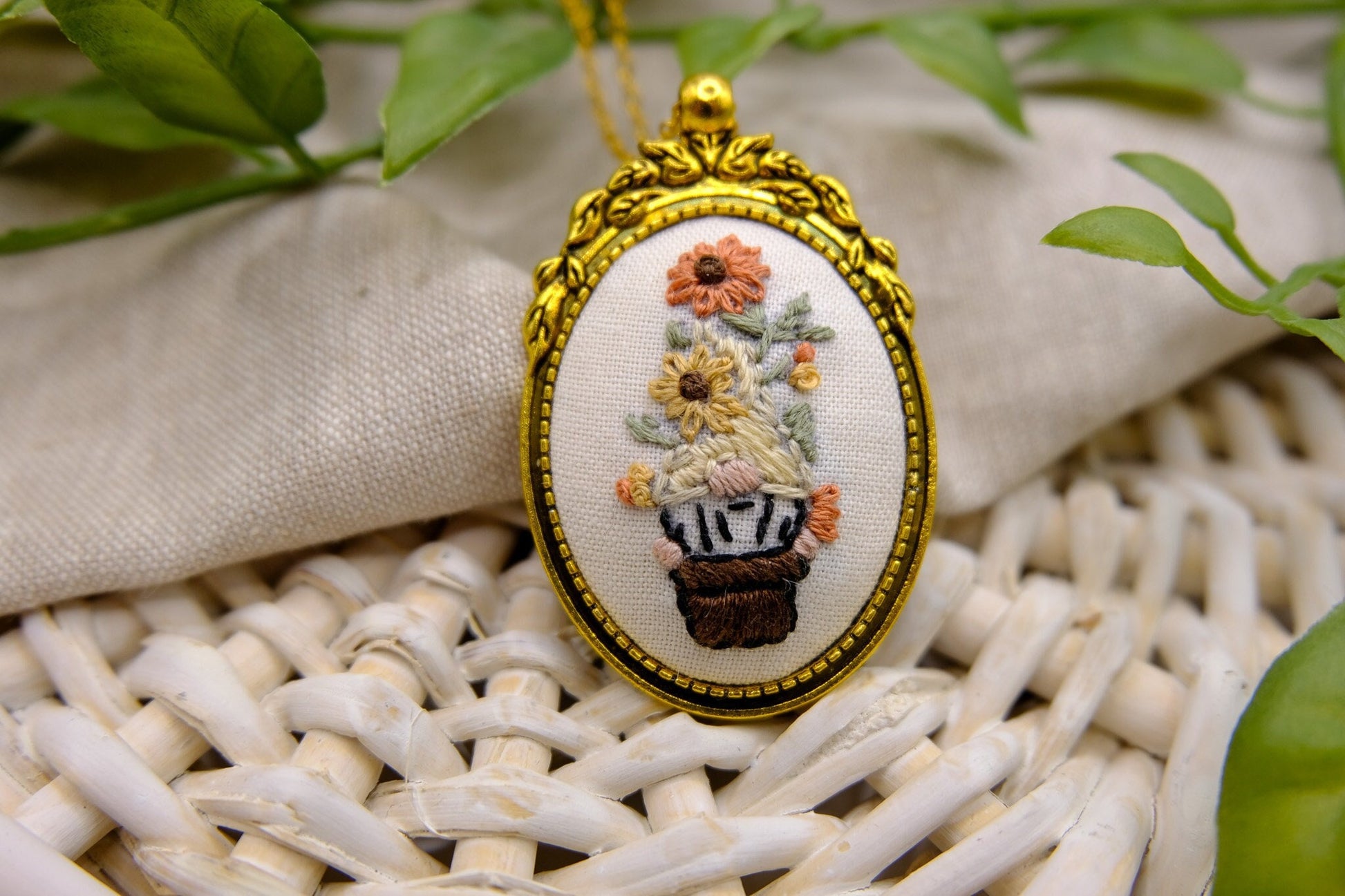 Embroidered pendant in a gold bezel featuring a gnome in a flower pot with flowers on his head