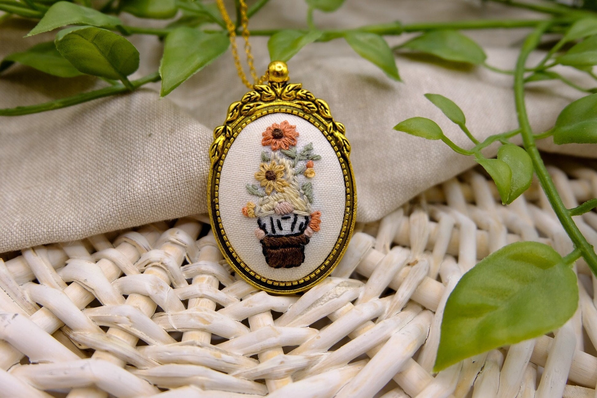 Embroidered pendant in a gold bezel featuring a gnome in a flower pot with flowers on his head