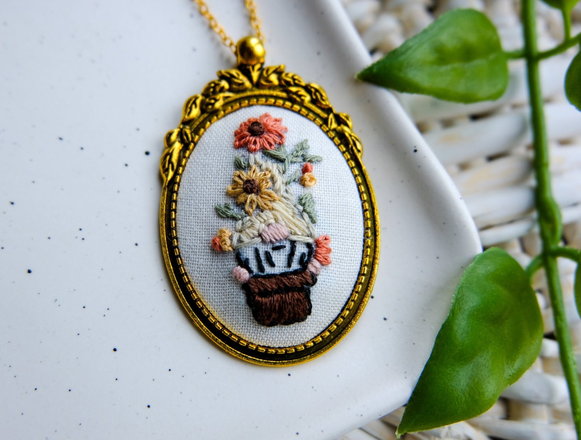 Embroidered pendant in a gold bezel featuring a gnome in a flower pot with flowers on his head